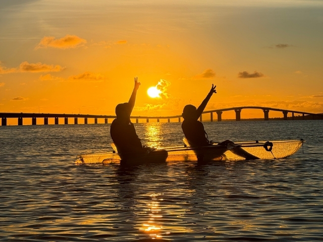 宮古島の伊良部大橋と夕日をバックに、クリアカヤックやSUPを楽しむ人々のシルエット