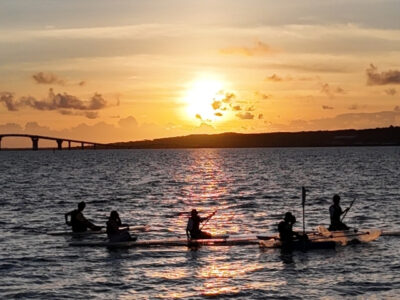 宮古島の伊良部大橋と夕日をバックに、クリアカヤックやSUPを楽しむ人々のシルエット